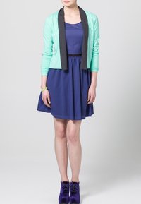 Mint-vihreä takki mustilla läpyillä sinisen mekon päällä. Mallilla on violetit nilkkurit. Sujuva kangas ja löysä istuvuus, jossa on elastinen vyötärö.