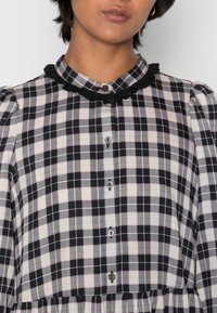 Černobílá kostkovaná blůza s volánovým černým límcem a knoflíkovým zapínáním. Má měkkou látku a nabírané rukávy.