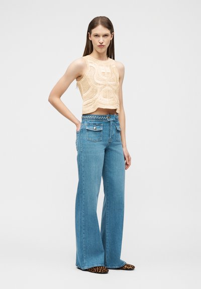 Femme portant un haut court beige en crochet sans manches, un jean large taille haute bleu avec poches sur la poitrine, et des ballerines à imprimé léopard, posant devant un fond blanc.