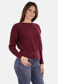 Maglione a maglia borgogna con texture a coste, maniche lunghe e un vita aderente, indossato con jeans blu su uno sfondo semplice.