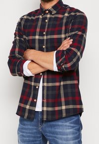 Koszula flanelowa w granatowo-czerwono-beżową kratę. Ma kołnierzyk na guziki oraz długie rękawy z podwiniętymi mankietami.