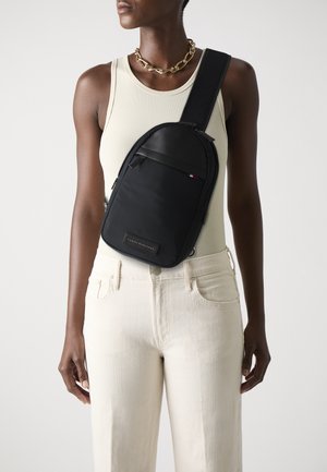 Schwarze Umhängetasche aus glattem Stoff, mit einem Hauptfach mit Reißverschluss, einer Vordertasche und einem Logopatch am Boden.