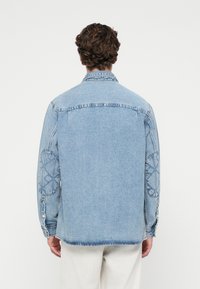 Jaqueta de ganga azul clara com fechos de botão, apresentando remendos bordados nos cotovelos e um corte descontraído. Detalhes de costura visíveis e um colarinho clássico.