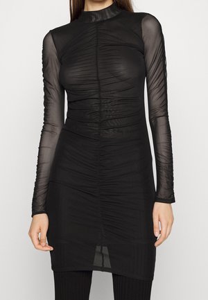 Kobieta w dopasowanej czarnej sukience z siateczki z marszczonymi detalami i długimi przezroczystymi rękawami, stojąca na jednolitym tle.