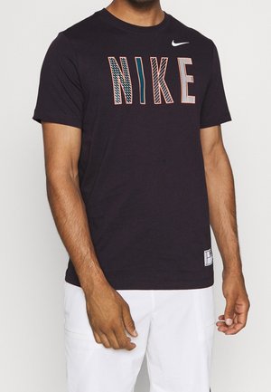 Mężczyzna w czarnej koszulce Nike z wzorzystym napisem NIKE i białych szortach, stojący ze swobodnie opuszczonymi ramionami.