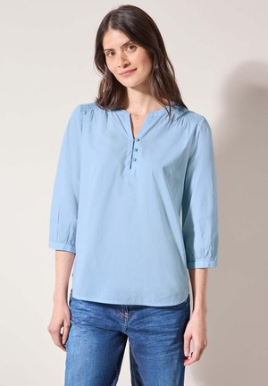 Blouse bleu clair à manches longues avec une patte de boutonnage à mi-hauteur, des détails froncés aux épaules et une coupe décontractée. Texture de tissu lisse.
