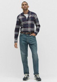 Koszula w kratę w kolorze granatowym i białym, nałożona na białą koszulkę, w zestawieniu ze średnimi niebieskimi dżinsami oraz czarnymi trampkami, stojąca na jednolitym tle.