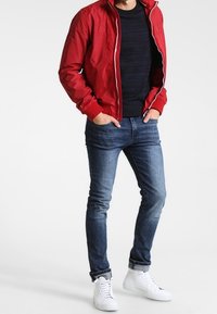 Red nylon bomber jacket with zipper, black textured shirt, blue jeans, and white sneakers. Casual outfit with a fitted design.