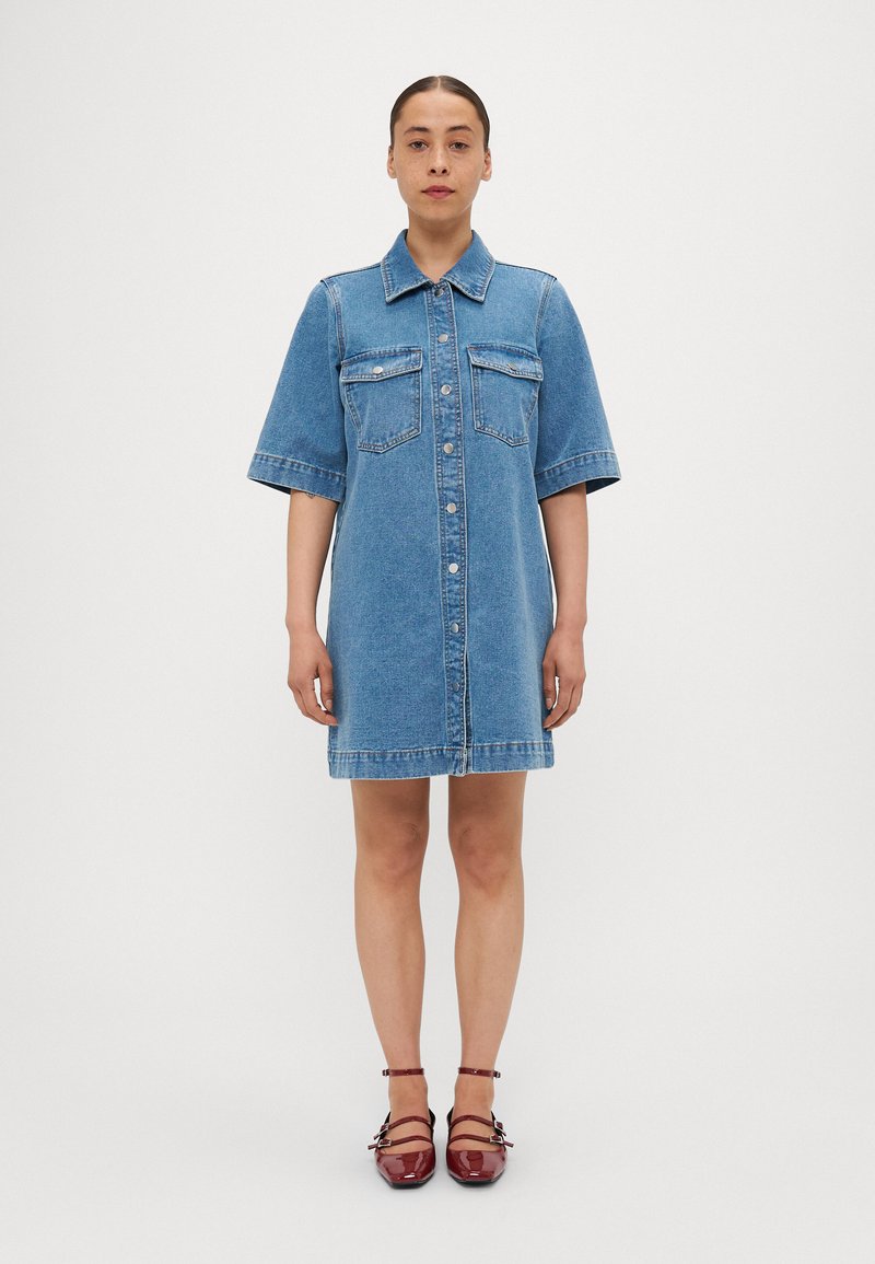 Jeanshemdkleid in Hellblau mit kurzen Ärmeln, Knopfverschluss, zwei Brusttaschen und geradem Schnitt, kombiniert mit roten Riemchen-Schuhen.