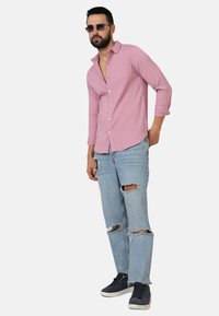 Camicia a maniche lunghe a strisce rosa con chiusura a bottoni, abbinata a jeans chiari strappati. Le scarpe sono sneakers nere.