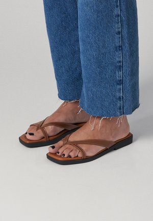 Pies con sandalias de cuero marrón con tiras cruzadas y esmalte de uñas negro, combinadas con vaqueros azules con dobladillo deshilachado sobre un fondo claro.