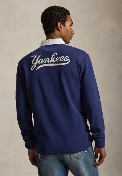 Férfi sötétkék hosszú ujjú ingben, fehér gallérral és a "Yankees" felirattal a háton, kék farmerrel párosítva.