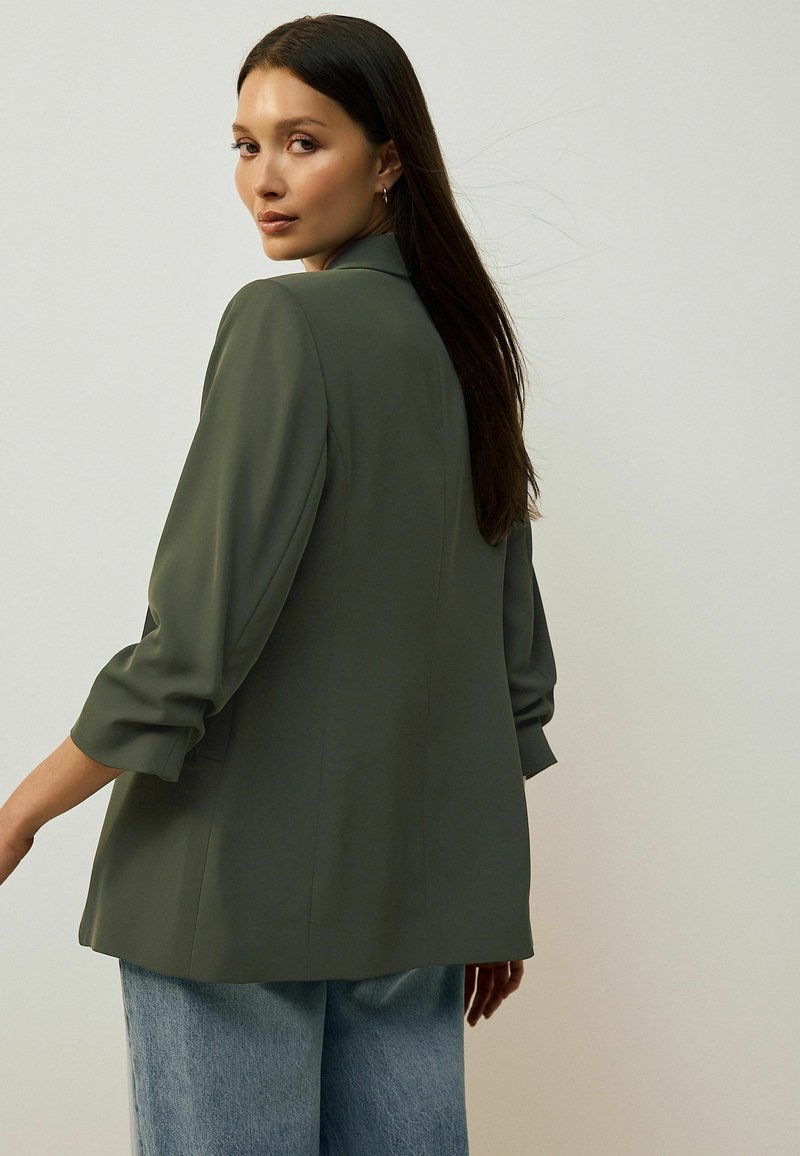 Olive green tailored blazer with three-quarter sleeves, soft texture, and a relaxed fit, paired with light blue wide-leg jeans.