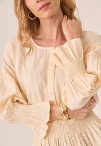 Blusa color crema con botones, tejido texturizado y mangas abullonadas. Se lleva una pulsera dorada brillante en la muñeca.