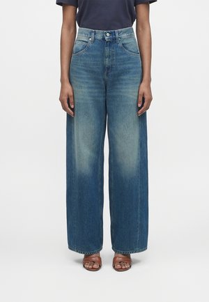 Femeie purtând blugi albaștri cu talie înaltă și croială largă, cu detalii șterse, asortați cu sandale maro cu vârf deschis și un top întunecat cu mâneci scurte.