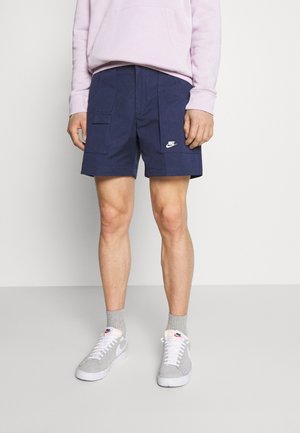 Shorts cargo bleu marine avec poches latérales, arborant un logo Nike blanc. Portés avec un sweat à capuche rose et des chaussettes grises, assortis à des baskets grises.