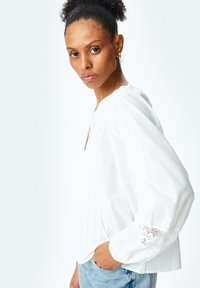 Hvid bluse lavet af letvægtigt stof, med V-hals, lange puffede ærmer og blonde detaljer på ærmeenderne. Casual pasform.
