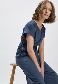 Robe en coton bleu à manches courtes et décolleté en V, dotée d'une taille cintrée et d'une silhouette fluide, présentée sur un tabouret contre un fond clair.