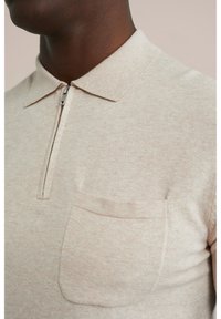 Beige polo shirt made of textured fabric with a zip collar and a front pocket. The collar is structured and the fabric has a subtle pattern.