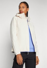 Giacca leggera con cappuccio, zip frontale, polsini elasticizzati e due tasche laterali. Indossata sopra una felpa blu e pantaloni scuri.
