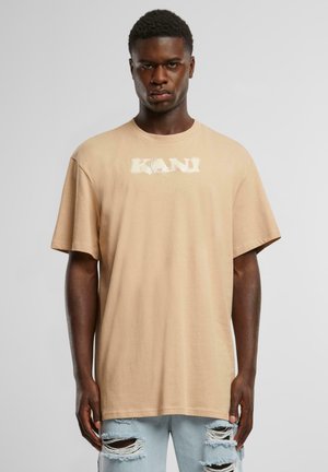 Beige bomull t-skjorte med korte ermer, med en hvit "KANI" logo på forsiden. Kombinert med slitte lyseblå jeans.