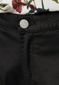 Svarta byxor med knappstängning och silverdetalj, sydda sömmar och mjuk textur. Delvis synlig blommig topp i bakgrunden.