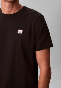 Mustast puuvillast T-särk ümarseljalõikega, vasakul rinnal väike valge logomärk. Lühikesed varrukad ja sile tekstuur.