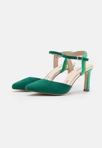 Zelené semišové lodičky na vysokém podpatku se špičatou špičkou a nártkovým páskem. Podpatek je štíhlý a béžový, s kontrastním zeleným detailem na pásku.