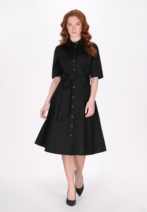 Schwarzes Hemdblusenkleid mit kurzen Ärmeln und tailliertem Bund. Fließender Rock und glatter Stoff. Getragen mit schwarzen spitzen Pumps.