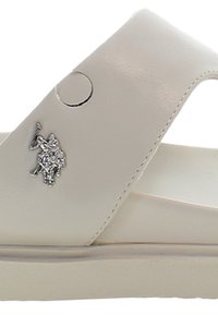 Beige syntetisk slidesandal med ett brett rem, cirkulär utskärning och metallisk logotypdetalj. Platt sula och slät yta.