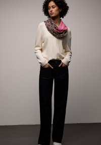 Femme aux cheveux bouclés portant un pull crème, une écharpe imprimée léopard rose et marron, un jean large foncé et des baskets blanches, les mains dans les poches.