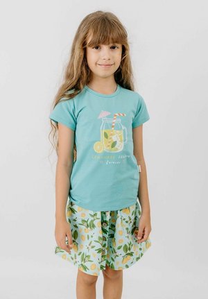 Young girl with long hair wearing a blue t-shirt with lemonade graphic and a lemon-patterned skirt, standing against a plain background.