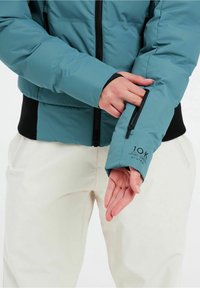 Blauer Puffer-Jacke mit figurbetontem Design, mit Reißverschlusstasche am Ärmel und schwarzen gerippten Bündchen. Das Material wirkt glatt und leicht glänzend.