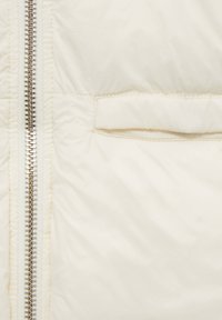 Pufferjacke in Cremeton, mit einem silbernen Reißverschluss, gestepptem Muster und einer kleinen Tasche auf der linken Seite.