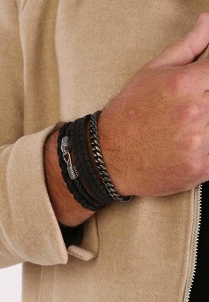 Vier armbanden zijn op een pols gestapeld: twee zwarte gevlochten leer, één zwarte gedraaide metalen ketting en één zilveren sluiting met een gestructureerd detail.