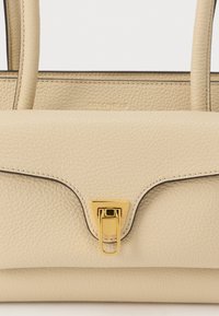 Beige leren handtas met een gladde textuur, voorzien van een goudkleurige sluiting en contrasterende stiksels. Dubbele bovenhandvatten met een subtiele logo-embossing.