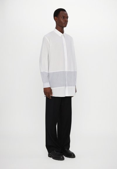 Chemise blanche à manches longues avec des détails à rayures horizontales bleues au niveau de la taille et des poignets. Associée à un pantalon noir à jambe large et des chaussures noires.