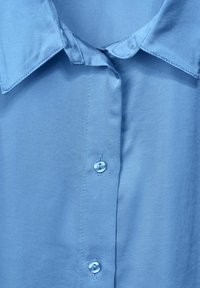 Camicia azzurra chiara con bottoni, realizzata in tessuto morbido, con colletto a punta e bottoni di plastica trasparente lungo la parte anteriore.