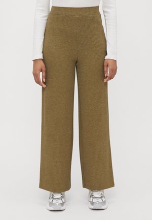 Vidbenade, ribbade byxor i olivgrön stickad tyg. Har en elastisk midja. Matchas med silvriga träningsskor.