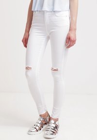 Vita smala jeans med hög midja, med två slitna knäsprickor, tillsammans med silver gladiatorsandaler. Slät textur och kroppsnära design.