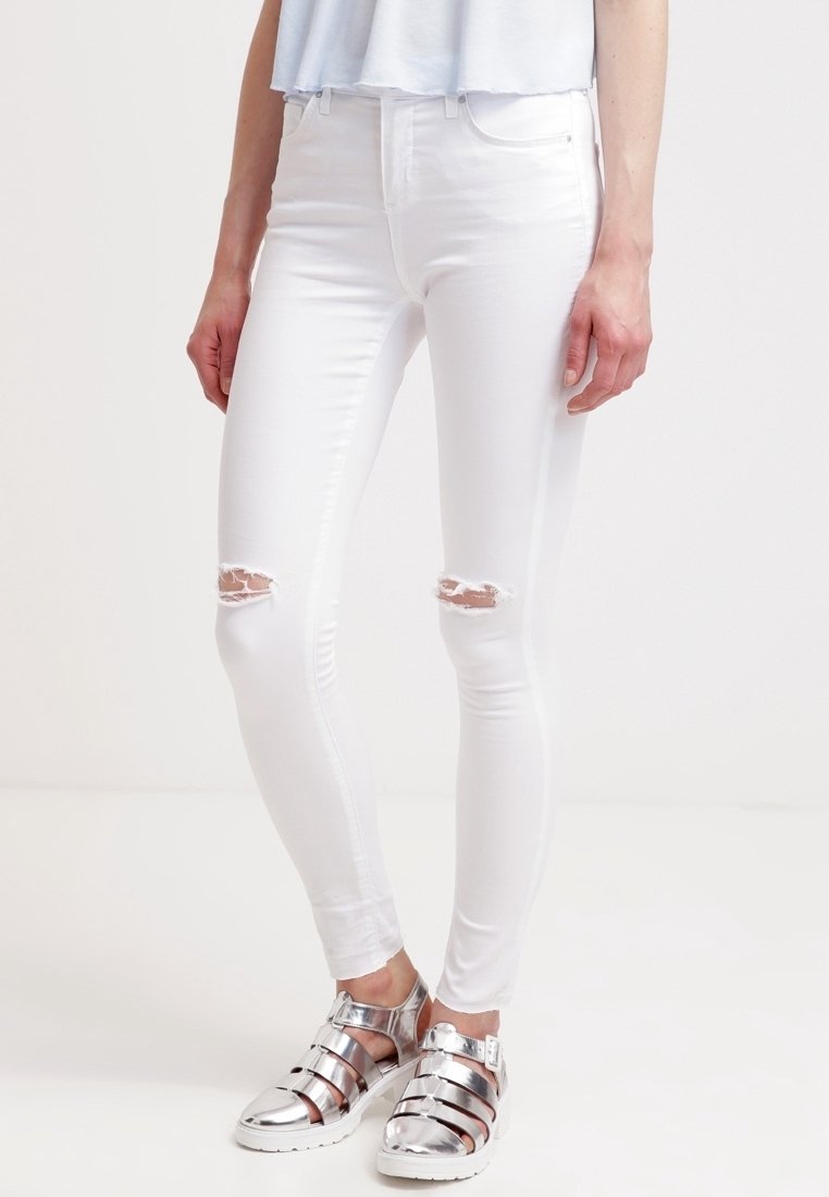 Vita smala jeans med hög midja, med två slitna knäsprickor, tillsammans med silver gladiatorsandaler. Slät textur och kroppsnära design.