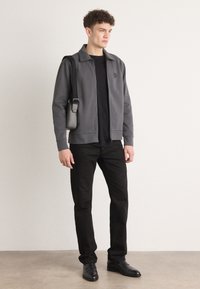 Giacca grigia con zip e colletto, T-shirt nera sotto, pantaloni neri e scarpe nere; modello che tiene una piccola borsa a tracolla nera.