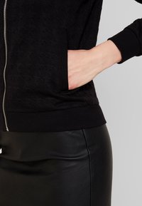 Svart zip-up jacka med texturerad tyg och ribbade ärmar. Har sidofickor och ett figursytt midjeband, ihop med en svart konstläderkjol.
