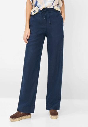 Vrouw die marineblauwe wijde broek met een taillekoord draagt, bruine espadrilleschoenen en een licht patroon top.