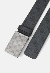 Sort mønstret læderbælte med en rektangulær sølvmetalsplade, der har et geometrisk design og prægede logo.