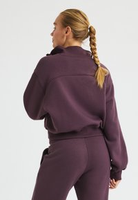 Lila sweatshirt med hög krage och ribbad mudd, matchande byxor. Tyget verkar mjukt och strukturerat, lämpligt för vardagsbruk.