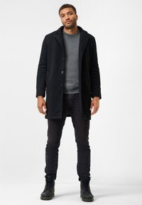 Sort frakke med en tekstureret finish, der har spidse klapper, knaplukning og sidelommer, båret over en grå crew-neck sweater og mørke bukser.