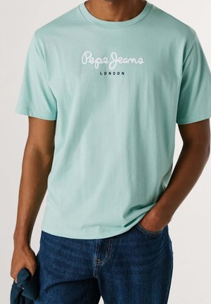 Mann trägt ein hellgrünes Pepe Jeans London T-Shirt mit kurzen Ärmeln und dunkelblaue Jeans, Hand in der Tasche und hält Stoff in der anderen Hand.