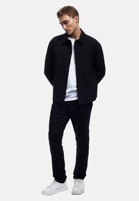 Svart knäppt jacka med två framfickor, lager på en vit t-shirt och matchad med svarta byxor och vita sneakers.