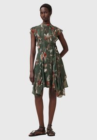 Floral-printed green dress with short ruffled sleeves, pleated design, and asymmetrical hem. Paired with black sandals.